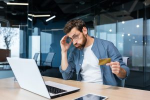 Cheated and disappointed businessman working inside office, man holding bank credit card and trying